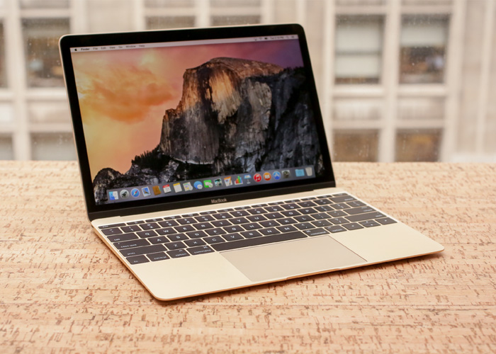 A gold 12 inch MacBook sits on a cork desk with a window in the background with another building across the street