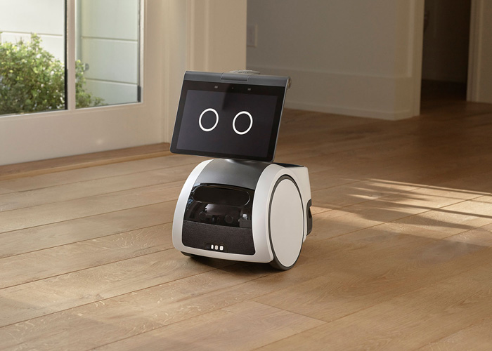 The white Amazon Astro household robot on wheels, displaying two large digital eyes on its screen-face, sitting on a hardwood floor.