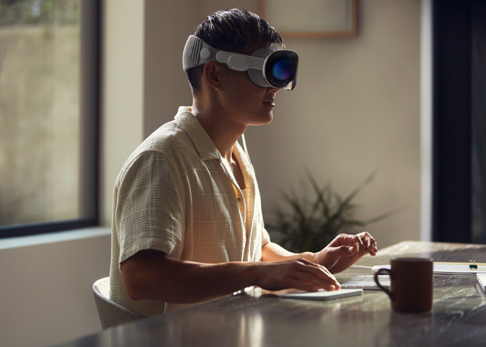 A person sitting at a desk in a modern with with an Apple Vision Pro strapped to their head. 