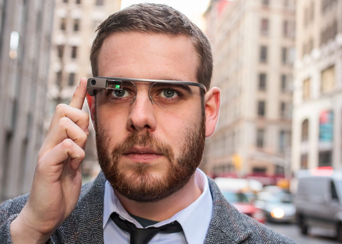 A man with a beard wearing Google Glass smart glasses on a city street, adjusting the side of the frame.
