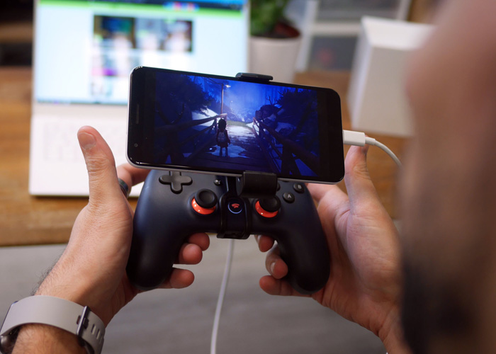 A person holding a Google Stadia gaming device with a white usb plug connected to a black phone.