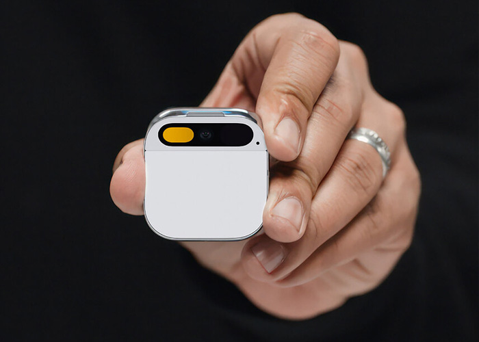 A hand holding the white Humane AI Pin, showing the touch surface and camera/projector array.