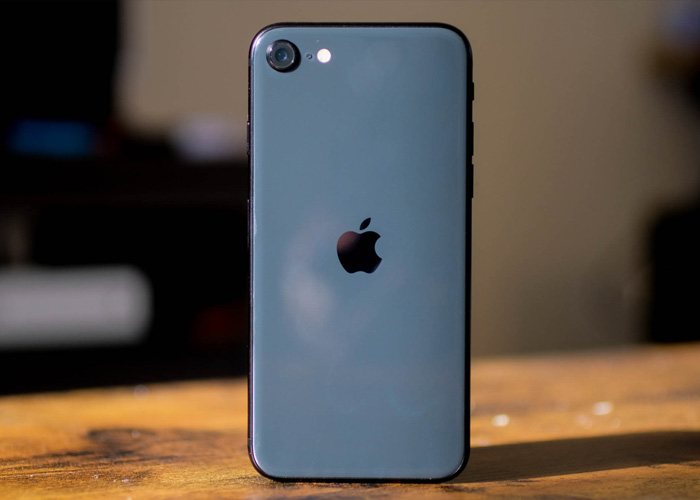 An iPhone SE sits on a wood desk with a blurry background in the distance. 