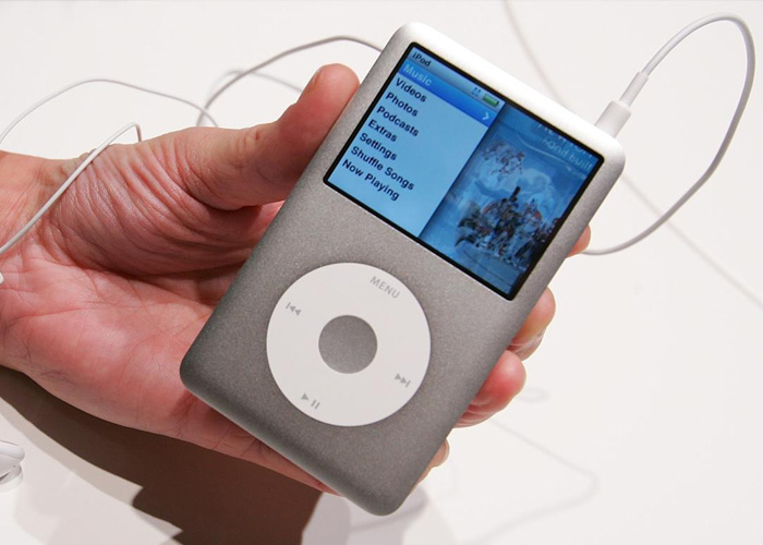 A person holds an iPod classic with video over a white desk. There are white headphones attached. 
