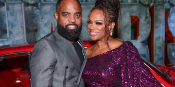 Todd Tucker wearing a grey blazer and Kandi Burruss wearing a sparkly purple sequin dress posing together in front of a classic red car.