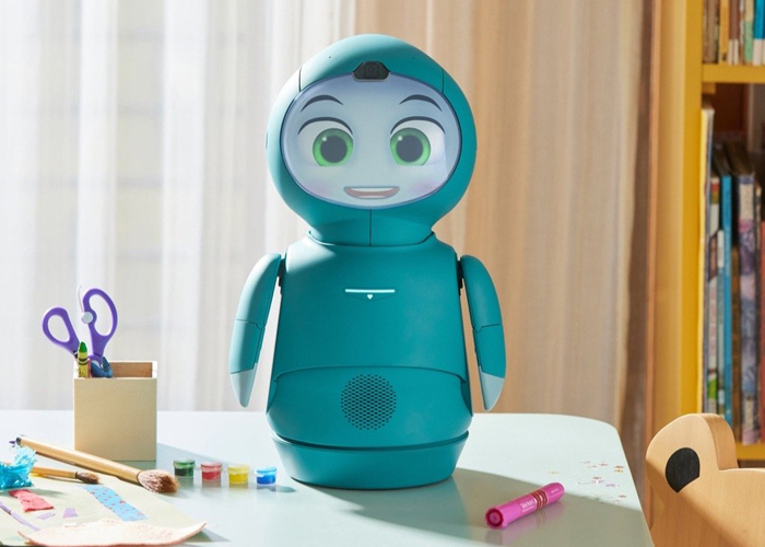 A Moxie AI robot sits on a desk with various desk objects like brushes and scissors. 