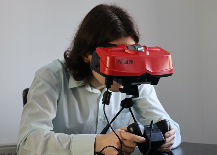 A person sitting at a table with a red Nintendo virtual boy with the controller in his hand.