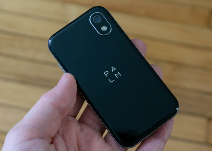 A hand holding a black palm phone with a wood floor in the background. 