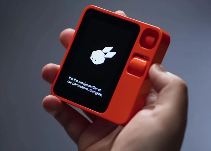 A hand holding the bright orange Rabbit R1 AI device, showing its small screen with a white rabbit logo.