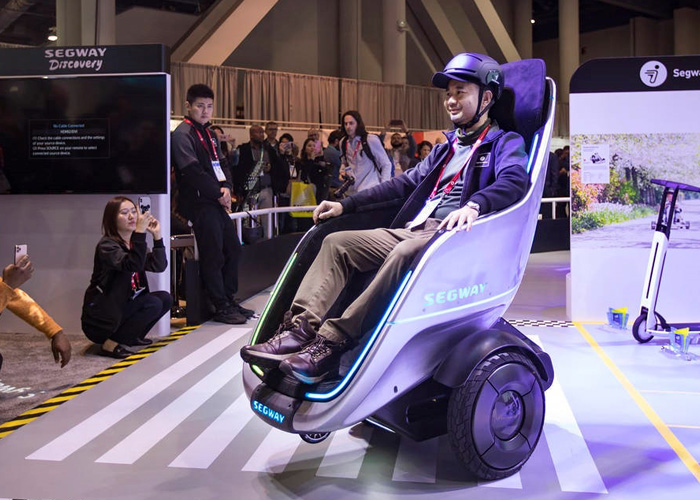 A presentation of a Sony S-Pod with a person with a helmet sitting inside of it with various onlookers in the background.