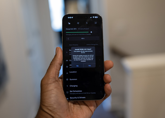 Alt-Text: A close-up, first-person view of a Black man’s hand holding an iPhone displaying the Tesla mobile app. A pop-up notification on the screen asks, 'Install 2026.2.9.1 Now?' and notes the update will take 55 minutes. The background shows a modern home interior softly blurred, emphasizing the convenience of initiating a Tesla Over-the-Air (OTA) software update remotely from a smartphone.