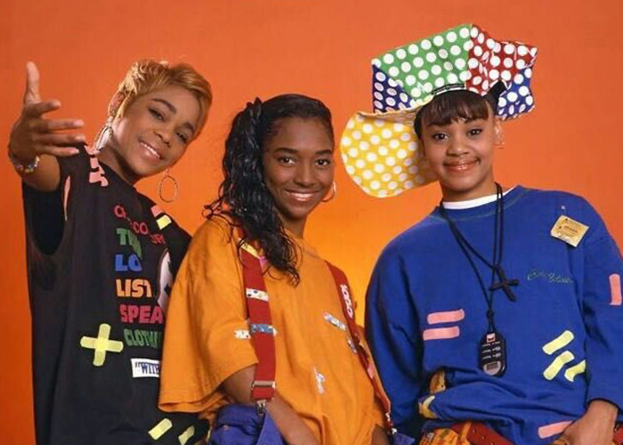 TLC (T-Boz, Chilli, and Lisa 'Left Eye' Lopes) in their signature colorful early 90s fashion, with Left Eye's multi-patterned hat, against a solid orange background.
