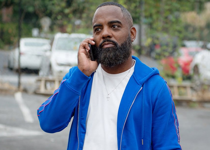 Todd Tucker in a blue zip up hoodie as he talks on the phone with cars in the background.